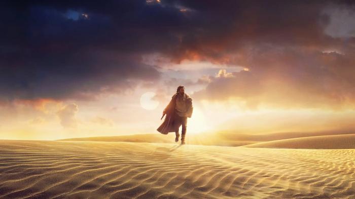 A still from the Disney+ show 'OBI-WAN KENOBI' showing Obi-Wan walking alone over sand dunes with dramatic dark clouds along with a bright late evening (or early morning) sun in the sky.