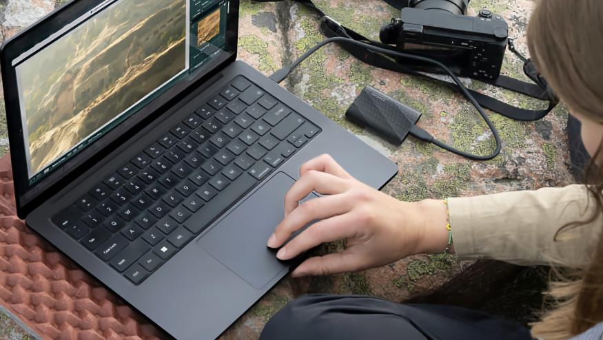 Lifestyle marketing photo of a person using a Windows laptop with a Samsung portable SSD plugged into it. A camera is also nearby, and it all sits on a green and orange surface.